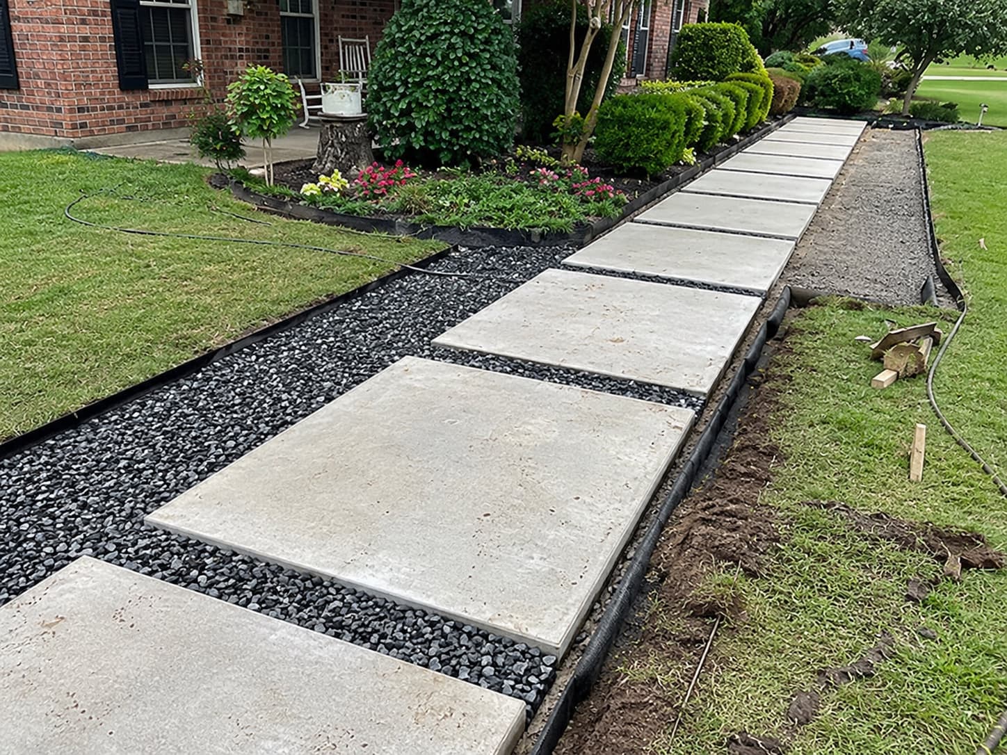 Large format concrete pavers set in black gravel with steel edging, forming a walkway alongside a brick home and its landscape beds